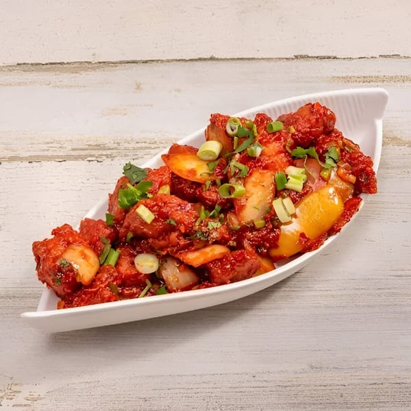 A white boat-shaped dish filled with vibrant red Indian chili chicken, garnished with green onions and cilantro.